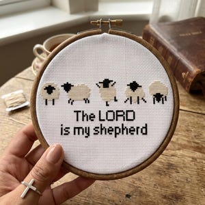 May include: A circular wooden embroidery hoop containing white fabric with a cross-stitch design of five sheep in various poses. Below the sheep, the text reads, "The LORD is my shepherd" in black thread. The hoop features a metal tightening screw.