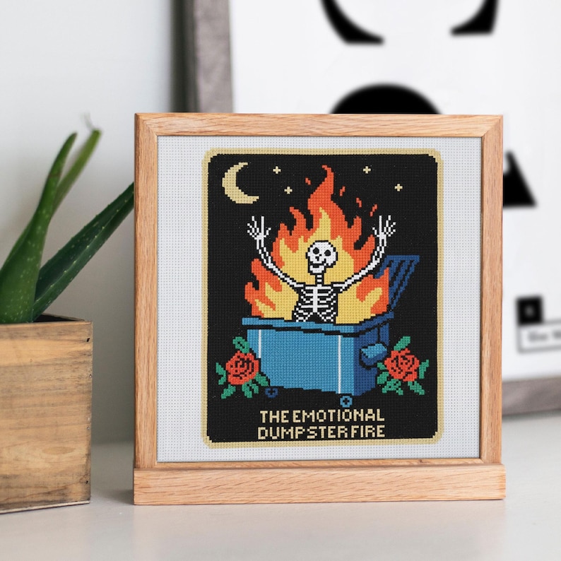 K&ouml;nnte beinhalten: Kreuzstich-Kunstwerk in einem Holzrahmen. Das Design zeigt ein Skelett in einem blauen M&uuml;llcontainer in Flammen, unter einem Mond und Sternen. Der Text lautet "THE EMOTIONAL DUMPSTER FIRE" mit roten Rosen unten.