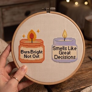 K&ouml;nnte beinhalten: Ein runder Stickrahmen aus Holz mit wei&szlig;em Stoff, der ein Kreuzstichmotiv von zwei Kerzen zeigt. Die orangefarbene Kerze tr&auml;gt den Text "Burn Bright Not Out" und die hellviolette Kerze den Text "Smells Like Great Decisions."