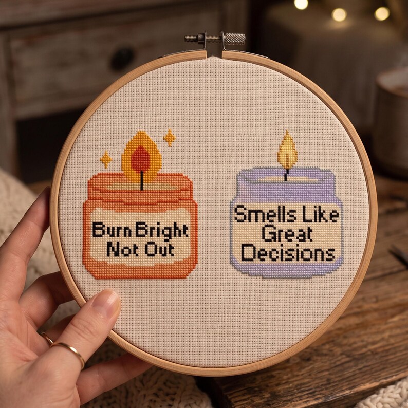 K&ouml;nnte beinhalten: Ein runder Stickrahmen aus Holz mit wei&szlig;em Stoff, der ein Kreuzstichmotiv von zwei Kerzen zeigt. Die orangefarbene Kerze tr&auml;gt den Text "Burn Bright Not Out" und die hellviolette Kerze den Text "Smells Like Great Decisions."