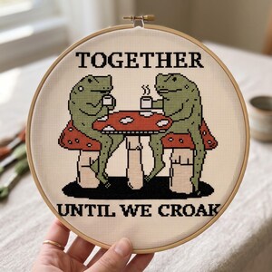 Pu&ograve; includere: Un telaio da ricamo circolare in legno contenente un disegno a punto croce di due rane verdi sedute a un tavolo a forma di fungo rosso e bianco mentre bevono da tazze bianche. Il testo TOGETHER UNTIL WE CROAK &egrave; ricamato in lettere nere.