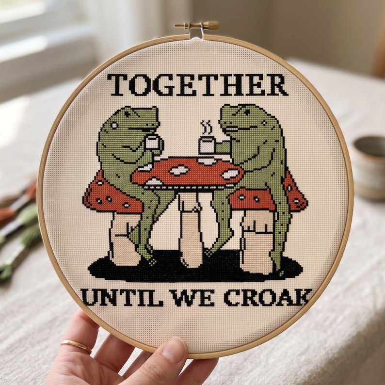 Pu&ograve; includere: Un telaio da ricamo circolare in legno contenente un disegno a punto croce di due rane verdi sedute a un tavolo a forma di fungo rosso e bianco mentre bevono da tazze bianche. Il testo TOGETHER UNTIL WE CROAK &egrave; ricamato in lettere nere.