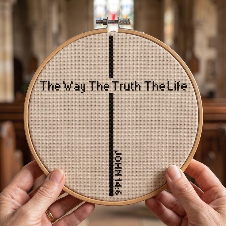 May include: A circular wooden embroidery hoop containing beige Aida fabric with black cross-stitched text reading The Way The Truth The Life and a vertical cross design with the text JOHN 14:6 positioned along the lower portion of the cross.