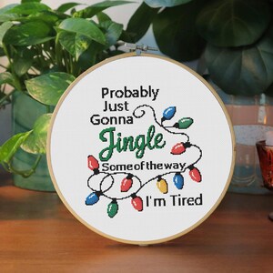 Może przedstawiać: Obręcz do haftu krzyżykowego z napisem "Probably Just Gonna Jingle Some of the way, I'm Tired" w kolorze czarnym i zielonym. Projekt zawiera sznur kolorowych lampek choinkowych. Obręcz jest umieszczona na tle zielonych roślin.