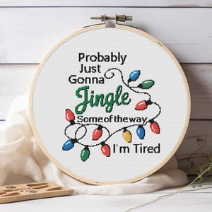 Może przedstawiać: Obręcz do haftu krzyżykowego z napisem "Probably Just Gonna Jingle Some of the way I'm Tired" w kolorze czarnym i zielonym. Projekt zawiera sznur kolorowych lampek choinkowych. Obręcz jest wykonana z drewna i umieszczona na białym tle.