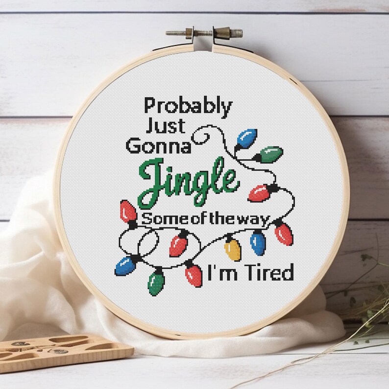 Może przedstawiać: Obręcz do haftu krzyżykowego z napisem "Probably Just Gonna Jingle Some of the way I'm Tired" w kolorze czarnym i zielonym. Projekt zawiera sznur kolorowych lampek choinkowych. Obręcz jest wykonana z drewna i umieszczona na białym tle.