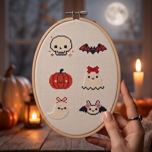 K&ouml;nnte beinhalten: Ein ovaler Stickrahmen aus Holz mit einem cremefarbenen Stoff, der sechs Halloween-Motive im Kreuzstich zeigt: einen wei&szlig;en Totenkopf, eine Fledermaus mit roten Fl&uuml;geln, einen orangefarbenen K&uuml;rbis, ein wei&szlig;es Gespenst mit roter Schleife und eine Fledermaus.