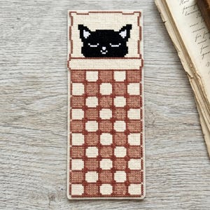 Pode incluir: Marcador de livros feito &agrave; m&atilde;o em ponto cruz com um gato preto a dormir. O gato est&aacute; numa almofada creme, coberto por um cobertor xadrez castanho e creme. O marcador de livros &eacute; retangular e feito de tecido.