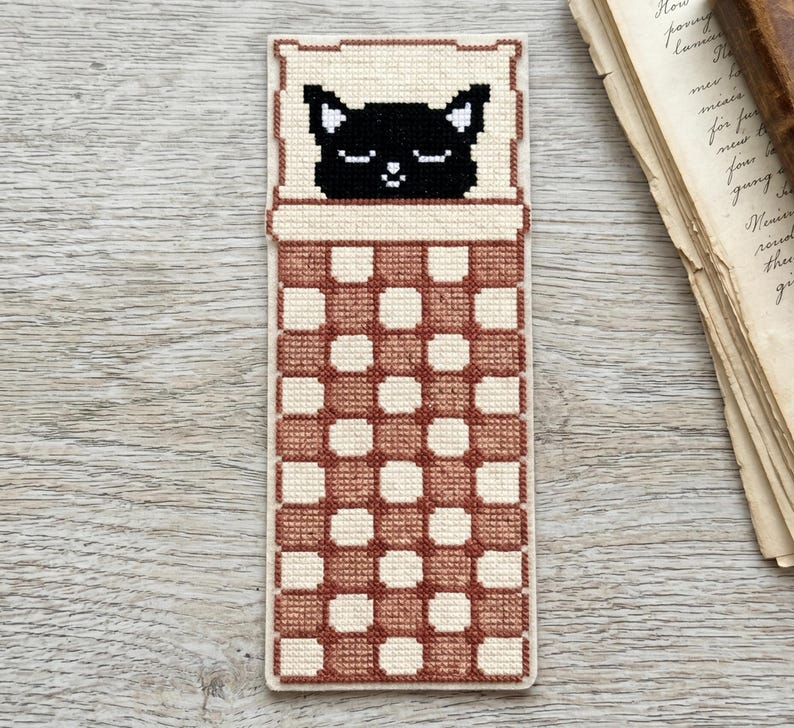 Pode incluir: Marcador de livros feito &agrave; m&atilde;o em ponto cruz com um gato preto a dormir. O gato est&aacute; numa almofada creme, coberto por um cobertor xadrez castanho e creme. O marcador de livros &eacute; retangular e feito de tecido.