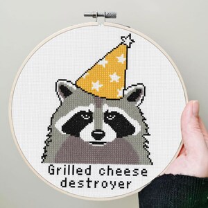 Op de afbeelding: Kruissteekborduurwerk in een houten hoepel met een wasbeer met een gele feesthoed met witte sterren. De tekst "Grilled cheese destroyer" staat eronder. Het ontwerp staat op witte stof.