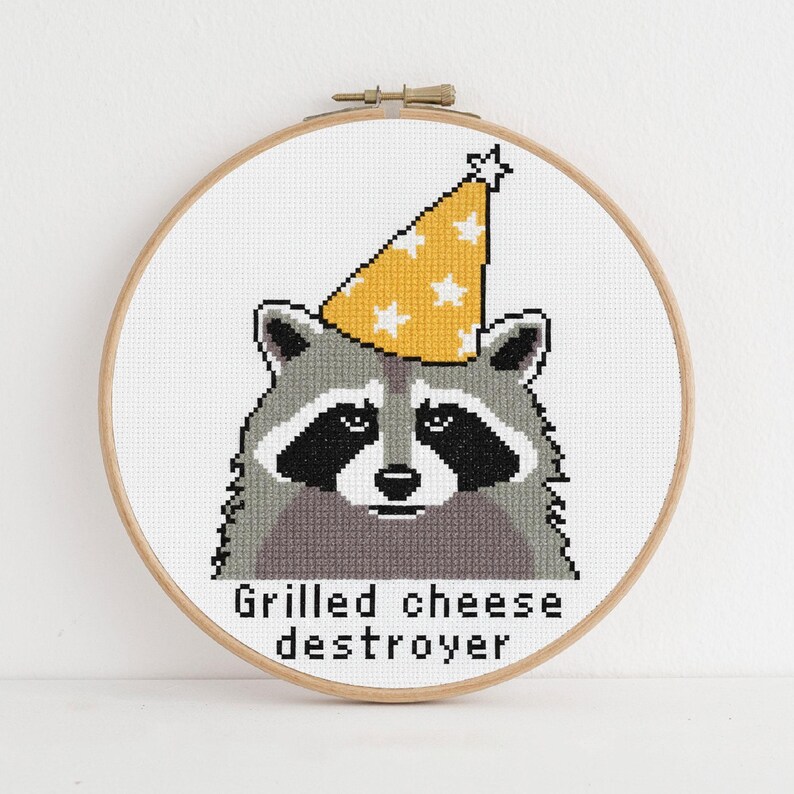 Op de afbeelding: Kruissteekborduurwerk van een wasbeer met een gele feesthoed met witte sterren. De wasbeer is grijs, zwart en wit. De tekst "Grilled cheese destroyer" staat eronder. De borduursel zit in een houten borduurring.