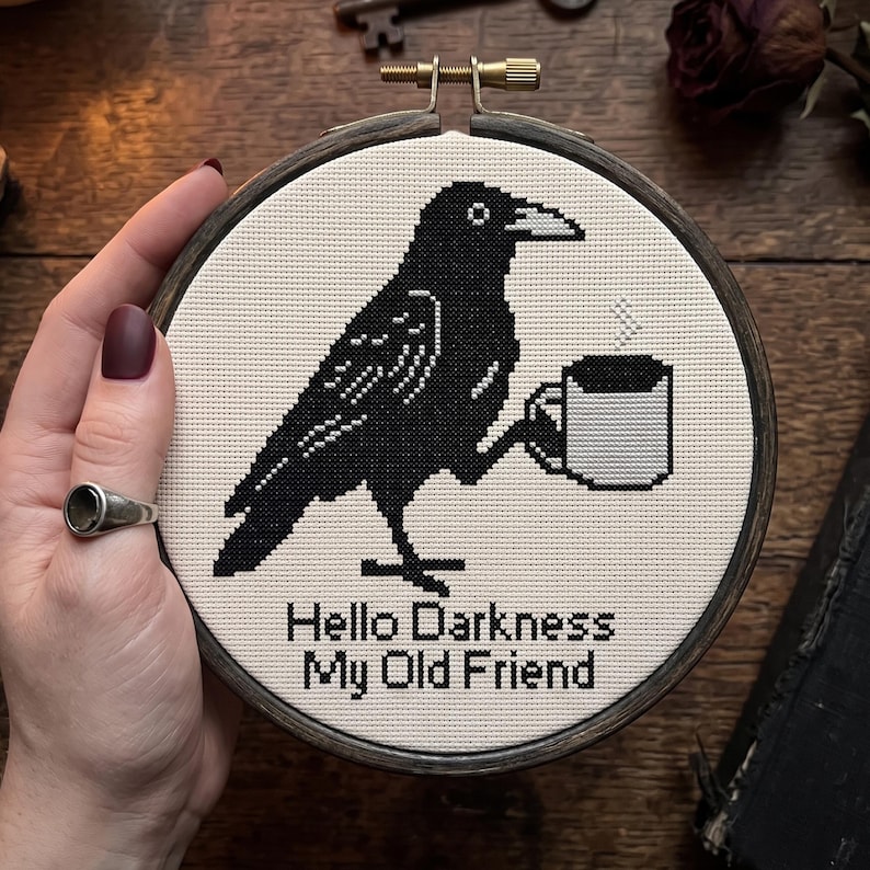 Op de afbeelding: Een ronde houten borduurring met een kruissteekontwerp op witte stof. Het ontwerp toont een zwarte kraai die een witte koffiemok met opstijgende stoom vasthoudt, en de zwarte tekst luidt: Hello Darkness My Old Friend.