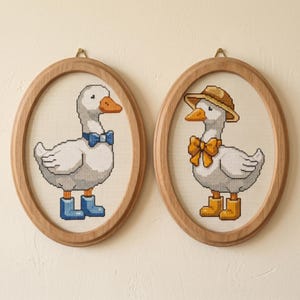 May include: Two framed cross-stitch pictures of geese. One goose has a blue bow tie and boots, the other a straw hat and yellow bow tie and boots. The oval frames are light wood, and the background is cream-coloured.