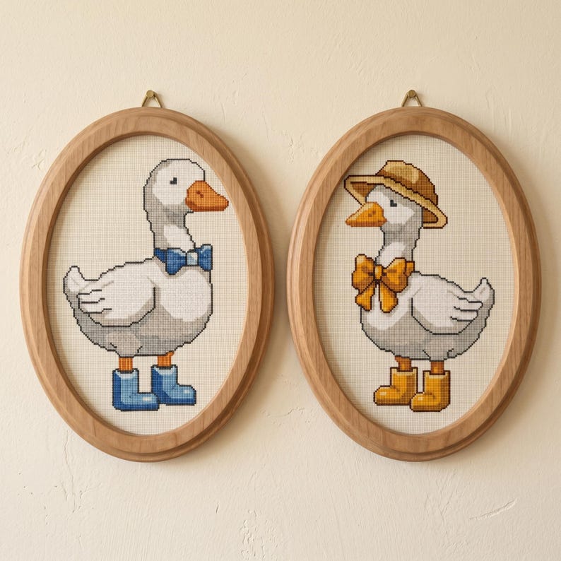 May include: Two framed cross-stitch pictures of geese. One goose has a blue bow tie and boots, the other a straw hat and yellow bow tie and boots. The oval frames are light wood, and the background is cream-coloured.