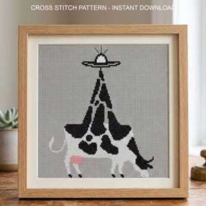 Pode incluir: Padr&atilde;o de ponto cruz emoldurado com uma vaca com manchas pretas e brancas, um &uacute;bere rosa e um OVNI acima. O texto "CROSS STITCH PATTERN - INSTANT DOWNLOAD" est&aacute; no topo. A obra de arte est&aacute; em uma moldura de madeira marrom clara.