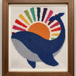 Op de afbeelding: Een ingelijst kruissteekpatroon met een blauwe walvis met een kleurrijk zonnestraalontwerp. Het kunstwerk staat tegen een witte achtergrond en is ingelijst in een donkerhouten frame. De tekst bovenaan luidt "CROSS STITCH PATTERN - INSTANT DOWNLOAD".