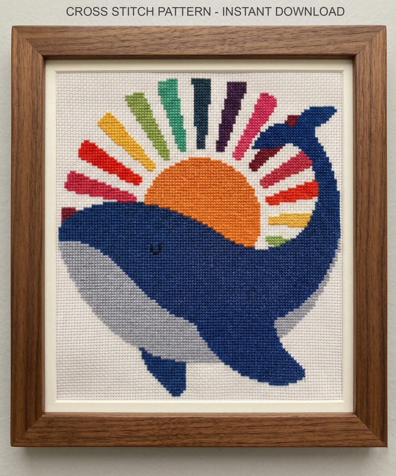 Op de afbeelding: Een ingelijst kruissteekpatroon met een blauwe walvis met een kleurrijk zonnestraalontwerp. Het kunstwerk staat tegen een witte achtergrond en is ingelijst in een donkerhouten frame. De tekst bovenaan luidt "CROSS STITCH PATTERN - INSTANT DOWNLOAD".