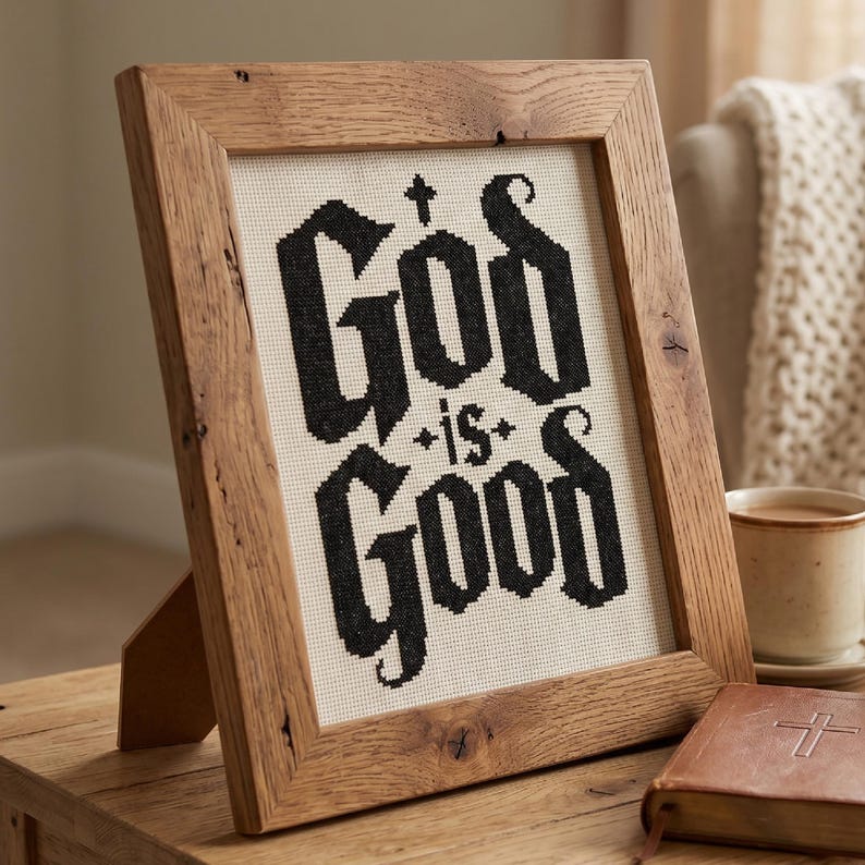 Peut inclure: Une broderie au point de croix encadr&eacute;e pr&eacute;sentant le texte noir "God is Good" sur un fond en tissu cr&egrave;me. L'&oelig;uvre est ins&eacute;r&eacute;e dans un cadre en bois brun clair rustique avec un grain visible, pos&eacute;e verticalement sur une surface plane.