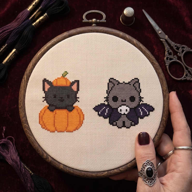 May include: A round wooden embroidery hoop containing white fabric with a cross-stitch design of a black cat sitting inside an orange pumpkin and a gray cat wearing a purple bat-wing cape with a small white skull accent on the chest.