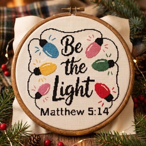 Może przedstawiać: Okrągły drewniany tamborek z białą tkaniną i wzorem wykonanym ściegiem krzyżykowym. Napis Be the Light Matthew 5:14 wyhaftowano czarną nicią, otaczając go sznurem kolorowych żar&oacute;wek w odcieniach niebieskim, ż&oacute;łtym, r&oacute;żowym, zielonym i czerwonym.