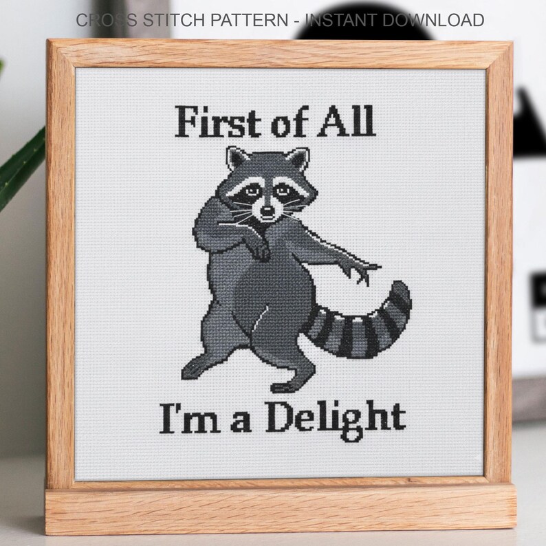 May include: Cross-stitch design in a wooden frame, depicting a raccoon with the text "First of All I'm a Delight". The raccoon is in shades of grey and black, set against a white background.