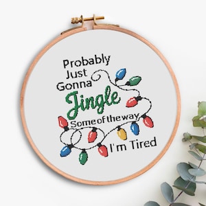 Może przedstawiać: Obręcz do haftu krzyżykowego z napisem "Probably Just Gonna Jingle Some of the way I'm Tired". Projekt zawiera sznur kolorowych lampek choinkowych.