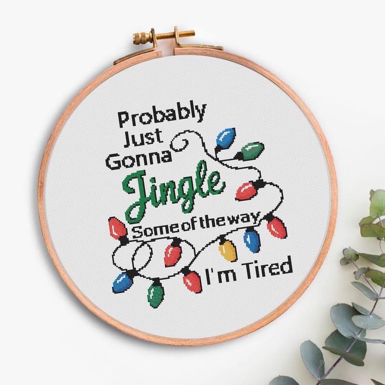 Może przedstawiać: Obręcz do haftu krzyżykowego z napisem "Probably Just Gonna Jingle Some of the way I'm Tired". Projekt zawiera sznur kolorowych lampek choinkowych.