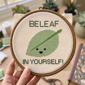 May include: A circular wooden embroidery hoop containing a cross-stitch design on white fabric. The design features a light green leaf with a smiling face and the dark green text "BELEAF IN YOURSELF!" stitched above and below the leaf.
