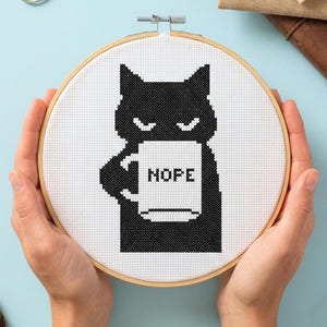 May include: A cross-stitch design in a wooden hoop shows a black cat holding a mug with the word "HOPE". The design is on white fabric. The text "CROSS STITCH PATTERN - INSTANT DOWNLOAD" is at the top. The hoop is held by hands.
