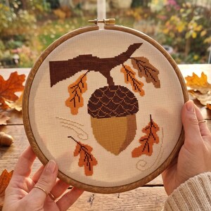 May include: A cross-stitch embroidery piece in a round wooden hoop featuring a large acorn hanging from a brown branch. The design includes several orange and brown oak leaves scattered around the acorn on a white fabric background.