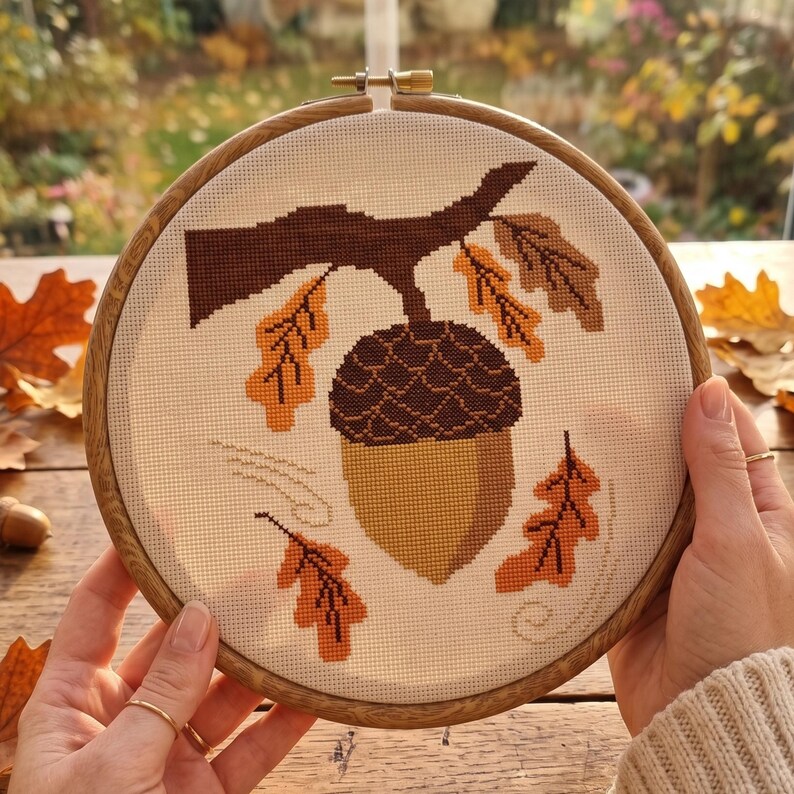 May include: A cross-stitch embroidery piece in a round wooden hoop featuring a large acorn hanging from a brown branch. The design includes several orange and brown oak leaves scattered around the acorn on a white fabric background.