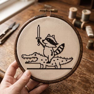 May include: A circular wooden embroidery hoop containing a piece of beige Aida fabric. The fabric features a black cross-stitch design of a raccoon holding a sword while riding on the back of a crocodile, rendered in a pixelated art style.
