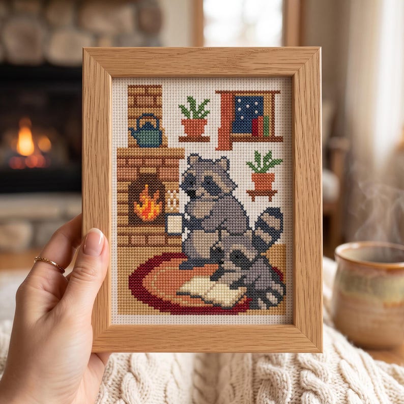 Puede incluir: Un bordado en punto de cruz enmarcado que muestra dos mapaches grises en una habitaci&oacute;n acogedora con chimenea de ladrillo, tetera azul, plantas en macetas y una ventana. Un mapache sostiene una taza blanca mientras el otro lee un libro abierto.