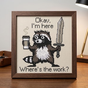 May include: Cross-stitch picture in a wooden frame showing a raccoon with a coffee cup and a sword. The text says "Okay, I'm here. Where's the work?" The raccoon is in brown, black, and white.