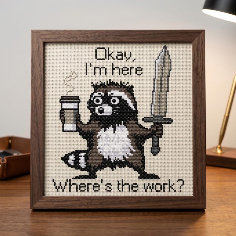 Puede incluir: Imagen de punto de cruz enmarcada en madera que muestra un mapache sosteniendo una taza de caf&eacute; y una espada. El texto dice: "Okay, I'm here. Where's the work?" El mapache es marr&oacute;n, negro y blanco.