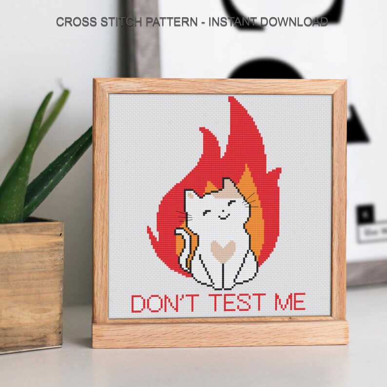 Pu&ograve; includere: Schema a punto croce in una cornice di legno. Il disegno presenta un gatto bianco con un marchio a forma di cuore, seduto di fronte a una fiamma rossa e arancione. Il testo "DON'T TEST ME" &egrave; sotto il gatto. La parte superiore della cornice recita "CROSS STITCH PATTERN - INSTANT DOWNLOAD".