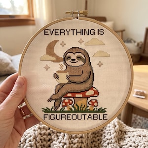 May include: A circular wooden embroidery hoop containing a cross-stitch design of a brown sloth sitting on a red and white spotted mushroom, holding a mug. The text reads EVERYTHING IS FIGUREOUTABLE, with a crescent moon and clouds in the background.