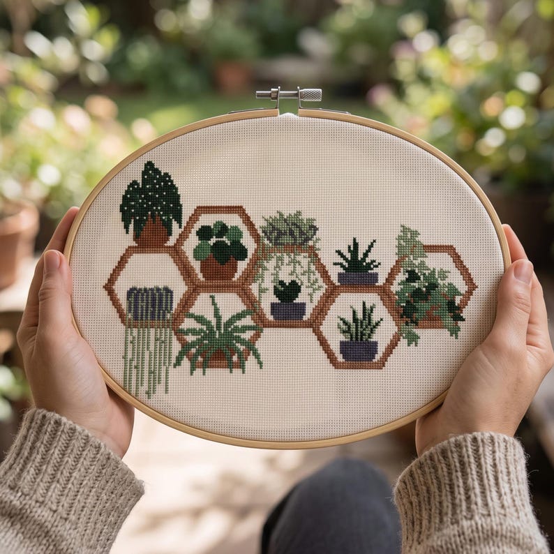 May include: An oval wooden embroidery hoop containing a cross-stitch pattern on white fabric. The design features various green potted plants arranged within interconnected brown hexagonal shelves, showcasing diverse foliage and plant shapes.