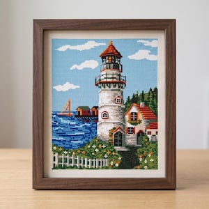 Peut inclure: Une broderie au point de croix encadr&eacute;e repr&eacute;sentant une sc&egrave;ne c&ocirc;ti&egrave;re avec un phare blanc, une maison au toit rouge, une mer bleue avec un voilier et une cl&ocirc;ture blanche. L'&oelig;uvre est ins&eacute;r&eacute;e dans un cadre en bois brun fonc&eacute; textur&eacute;.