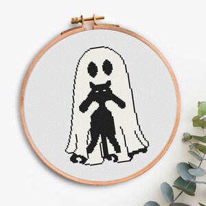 K&ouml;nnte beinhalten: Ein Kreuzstich-Stickrahmen mit einem Halloween-Design. Das Design zeigt eine schwarze Katze in einem wei&szlig;en Geisterkost&uuml;m. Die Augen und der Mund der Katze sind durch die Augenl&ouml;cher des Geistes sichtbar. Der Rahmen ist aus Holz.