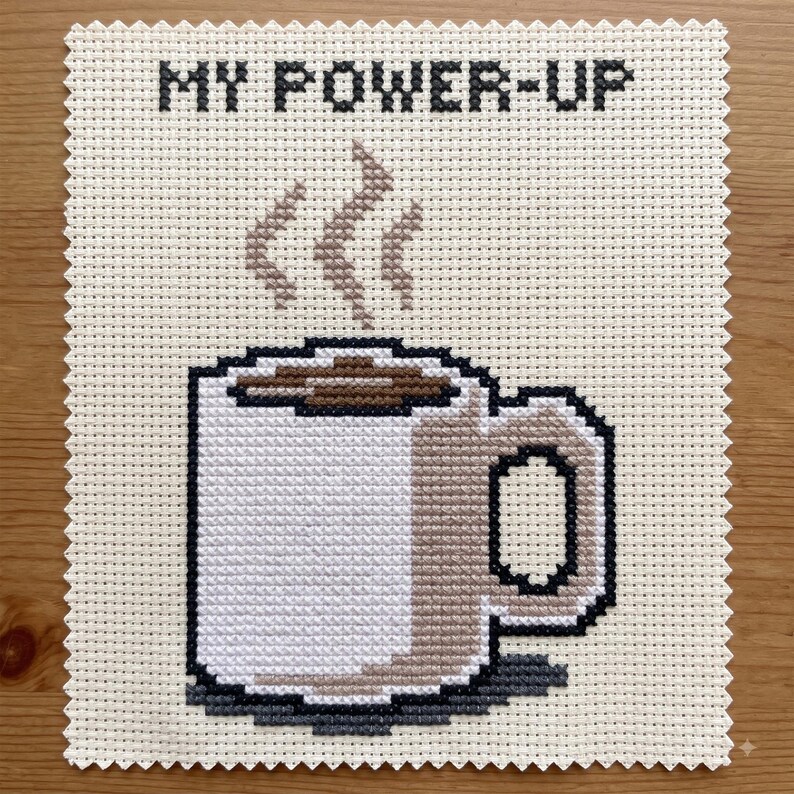 Puede incluir: Un bordado de punto de cruz sobre tela Aida color crema que muestra una taza de caf&eacute; blanca con l&iacute;quido marr&oacute;n y vapor elev&aacute;ndose. El texto MY POWER-UP est&aacute; bordado en letras negras de bloque en la parte superior. Los bordes tienen corte dentado.