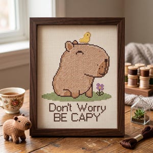 May include: A framed cross-stitch embroidery featuring a tan capybara with a small yellow bird on its head and a purple flower on green grass. The text below reads Don't Worry BE CAPY. The piece is enclosed in a dark brown wooden frame.