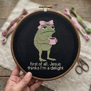 Puede incluir: Un bastidor de bordado circular de madera con tela negra y un sapo verde bordado en punto de cruz, con un lazo rosa y una taza rosa. El texto debajo del sapo dice, First of all, Jesus thinks I'm a delight, bordado en hilo de color blanco.