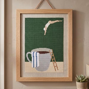 Puede incluir: Una pieza de arte en punto de cruz enmarcada con un dise&ntilde;o pixelado de una persona saltando a una gran taza de caf&eacute; gris. La taza se asienta sobre una superficie beige frente a un fondo verde oscuro, con una peque&ntilde;a escalera de madera y una toalla a rayas azules y blancas.