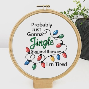 Może przedstawiać: Obręcz do haftu krzyżykowego z napisem "Probably Just Gonna Jingle Some of the way, I'm Tired." Projekt przedstawia sznur kolorowych lampek choinkowych w kolorach czerwonym, zielonym, niebieskim i ż&oacute;łtym. Obręcz jest wykonana z drewna i stoi na drewnianym stojaku.