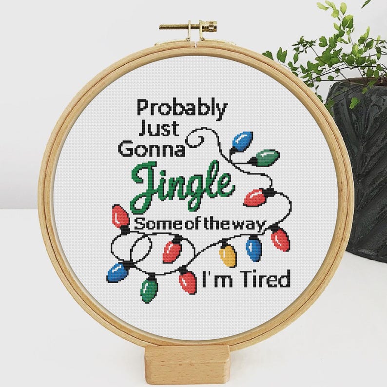 Może przedstawiać: Obręcz do haftu krzyżykowego z napisem "Probably Just Gonna Jingle Some of the way, I'm Tired." Projekt przedstawia sznur kolorowych lampek choinkowych w kolorach czerwonym, zielonym, niebieskim i ż&oacute;łtym. Obręcz jest wykonana z drewna i stoi na drewnianym stojaku.