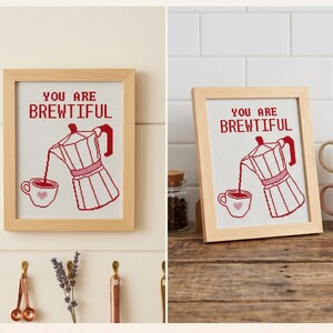 K&ouml;nnte beinhalten: Ein gerahmtes Kreuzstichbild mit einer roten Mokkakanne, die Kaffee in eine kleine wei&szlig;e Tasse mit einem rosa Herz gie&szlig;t. &Uuml;ber der Mokkakanne steht der Text YOU ARE BREWTIFUL in roten Gro&szlig;buchstaben. Das Kunstwerk befindet sich in einem hellen Holzrahmen.