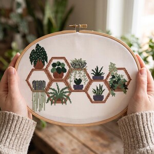 May include: An oval wooden embroidery hoop containing white Aida fabric with a cross-stitch pattern of various green potted plants arranged inside interconnected brown hexagonal shelves, featuring diverse foliage shapes and shades of green.