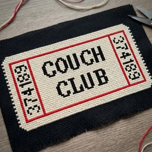 May include: A rectangular cross-stitch embroidery on black fabric featuring a cream-coloured ticket design with a red border. The text COUCH CLUB is stitched in black, flanked by the number 374189 on both sides in a vertical orientation.