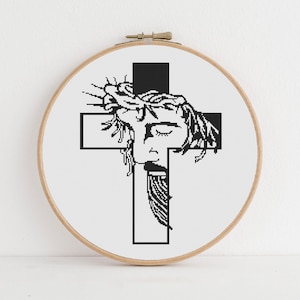 Puede incluir: Un bastidor de bordado circular de madera que contiene un patr&oacute;n de punto de cruz de una ilustraci&oacute;n gr&aacute;fica en blanco y negro. El dise&ntilde;o presenta un rostro estilizado con una corona de espinas sobre una cruz negra en fondo blanco.